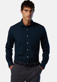 Camisa de manga larga azul marino con botones, con un cuello clásico, que presenta un pequeño logo y botones blancos, combinada con pantalones grises y un cinturón trenzado.