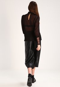Blouse noire transparente avec des poignets en dentelle, détail de dos en trou de serrure rond, associée à un pantalon culotte noir en faux cuir taille haute et des chaussures noires épaisses.