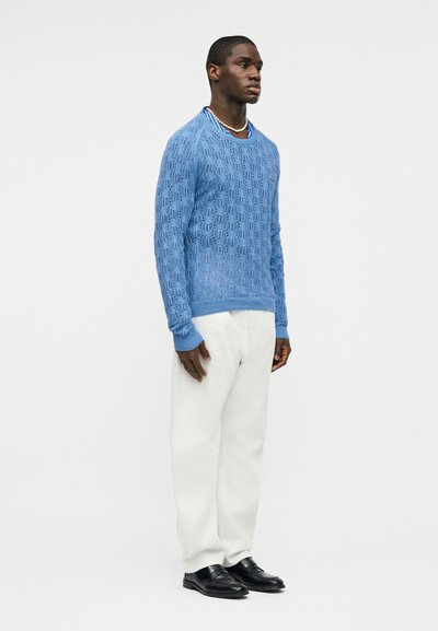 Homme debout portant un pull en maille bleu clair, un pantalon blanc, des mocassins noirs et un collier de perles, sur un fond blanc uni.