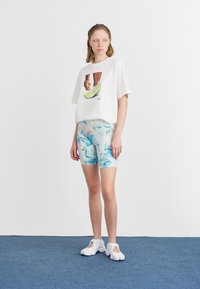 Fehér, cropped t-shirt grafikai mintával és kék, absztrakt mintázatú rövidnadrággal. A modellen fehér sportcipők vannak egy kék szőnyeg háttér előtt.