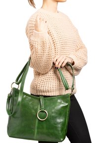 Borsa a spalla in pelle verde con una texture lucida, caratterizzata da un accento in oro rotondo e una tracolla rimovibile, tenuta da una persona.