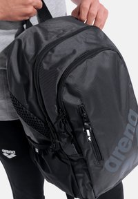 Zaino nero in nylon con spallacci regolabili, molteplici scomparti con cerniera e dettagli in rete. Presenta un grande logo sul lato.