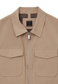 Giacca beige con collo a camicia, chiusura con zip e due tasche applicate. Realizzata in tessuto testurizzato, con dettagli di cucitura bianca.
