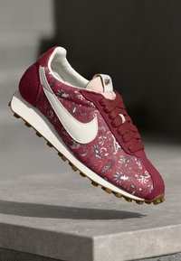 Crvene tenisice s uzorkom, bijeli Swoosh logo, gornji dio od mekog materijala, teksturirani bočni detalji i smeđi gumeni đon. Ima vezice i podstavljeni ovratnik.