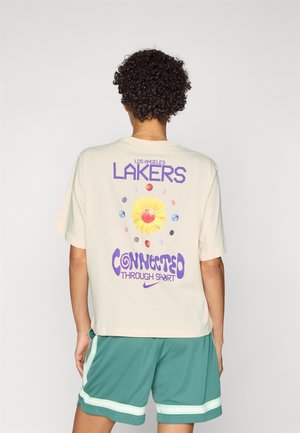 Personne aux cheveux courts et bouclés porte un t-shirt beige des Los Angeles Lakers avec un motif tournesol et basket-ball, short de sport vert avec des rayures blanches.