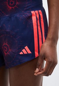 Κοντινό πλάνο σε σκούρο μπλε αθλητικό σορτς με κόκκινες αφηρημένες κυκλικές ρίγες, τρεις κάθετες κόκκινες ρίγες και κόκκινο λογότυπο Adidas στην πλευρά.