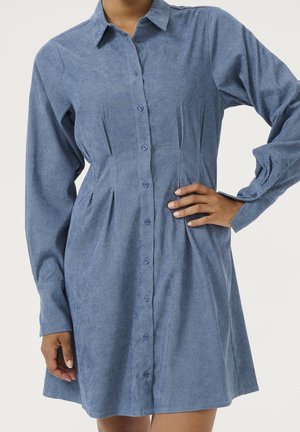 Kvinde iført blå manchesterkjole med knapper, lange ærmer, krave og plisserede detaljer i taljen, poserede med den ene hånd på hoften.