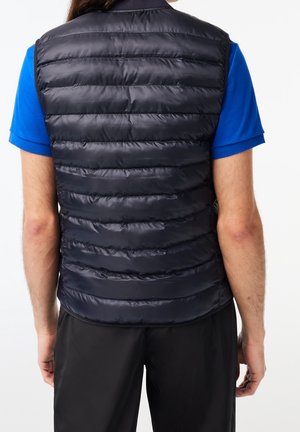 Smanicato imbottito navy con cuciture orizzontali, leggero e con finitura lucida. Indossato sopra una camicia blu, abbinato a pantaloni neri.