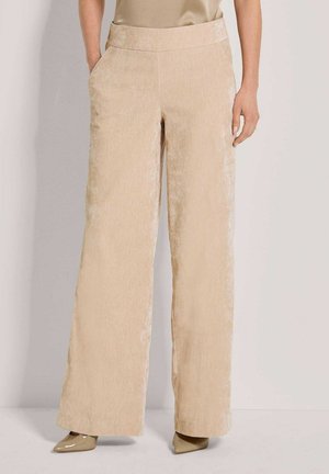 Femme portant un pantalon en velours beige taille haute, jambes larges, les mains dans les poches et des chaussures beiges à bout pointu sur un fond uni.