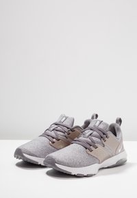 Un paio di scarpe da training Nike grigie con allacciatura, colli imbottiti e suole bianche su una superficie bianca contro uno sfondo grigio chiaro.