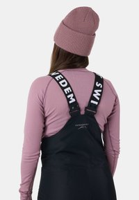Zwarte tuinbroeken met verstelbare banden met "SWEDMOUNT" branding, gecombineerd met een lichtroze geribbelde lange mouw shirt en een bijpassende gebreide beanie.