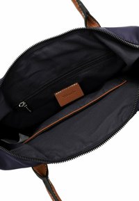 Interno della borsa in tessuto blu navy con accenti in pelle marrone. Presenta una tasca con zip e una texture liscia. La fodera è scura con un etichetta logo ricamata.