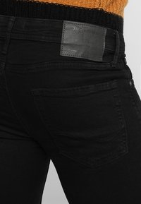 Musta farkkujen denimi, jossa sileä pinta, nahkainen merkki vyötärönauhassa ja yksittäinen takatasku, jossa on kaareva ompelukuvio.