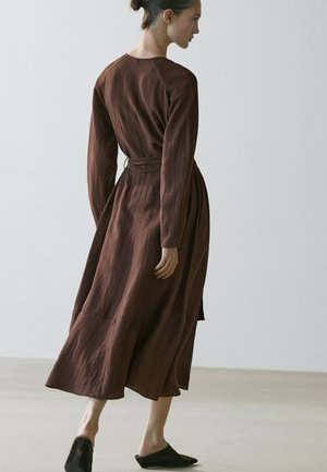 Kobieta w brązowej, długorękawowej sukience midi chodząca boso po jasnej podłodze na tle gładkiej, jasnej ściany.