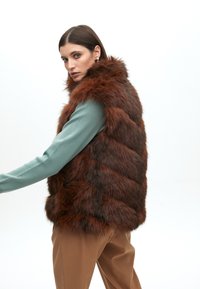 Hnědá vestu z umělé kožešiny s vysokým límcem, s prošívaným vzorem a bočními kapsami, nošenou přes tyrkysový svetr a hnědé kalhoty.