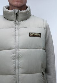 Gilet imbottito grigio chiaro con collo alto, dotato di una cerniera frontale e di una toppa logo nera e arancione. Il materiale ha una consistenza liscia e lucida.