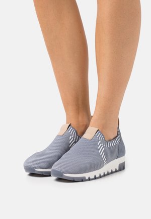 Legs wearing slip-on blue knit sneakers with white soles, standing on a white surface.