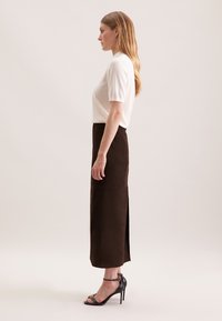 Top blanc à manches courtes associé à une jupe midi en daim marron foncé avec une fente sur le côté. Des sandales à talons aiguilles noires complètent la tenue.