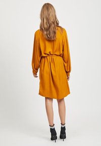Robe mi-longue couleur moutarde avec des manches longues, taille ceinturée et texture lisse. Bottines noires avec des rayures blanches et un talon haut.