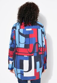 Camicia a righe geometriche multicolori con sezioni blu, rosse e bianche, vestibilità ampia, maniche lunghe e dettagli ai polsini.