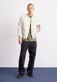 Giacca bomber bianca con accenti grigi, maglietta verde oliva, jeans scuri e scarpe chunky nere. Dettaglio della cerniera e portachiavi notevoli.