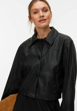 Femme portant une veste en cuir noire boutonnée avec un col, debout devant un fond blanc uni, face à l'appareil.