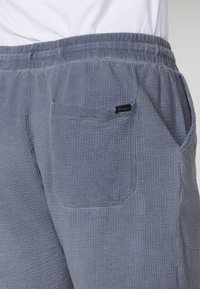 Ingrandimento di pantaloni in tessuto blu texturizzato con tasca posteriore e cintura elastica, indossati con una camicia bianca.