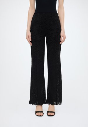 Μαύροι ψηλόμεσοι δαντέλα παντελόνια με καμπάνα στα πόδια, φορεμένοι με μαύρα πέδιλα με λουριά και ανοιχτά δάχτυλα, σε ένα πρόσωπο που στέκεται μπροστά σε λευκό φόντο.