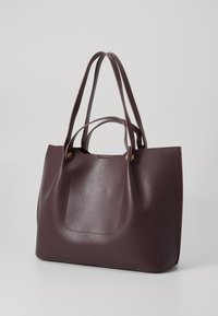 Sac cabas en cuir bordeaux avec deux longues poignées, surface texturée et poche avant plaquée. Présente des accents en métal doré.
