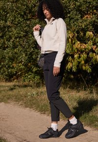 Veste en polaire de couleur crème, zippée, associée à un pantalon noir et des baskets foncées. La scène comprend de la verdure et un chemin en terre.
