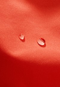 Tissu rouge avec une texture lisse, présentant deux gouttes d'eau claires reposant à la surface. Le tissu semble vibrant et brillant.