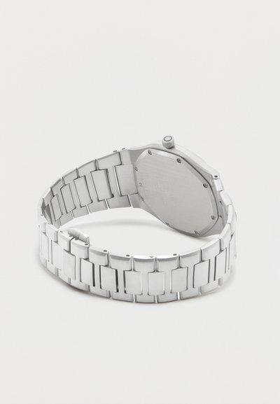 Ασημένιο μεταλλικό ρολόι χειρός με συνδεδεμένο λουράκι, εμφανιζόμενο από πίσω, δείχνοντας την κλειστή θήκη και το κουμπί ρύθμισης πάνω σε απλό φόντο.