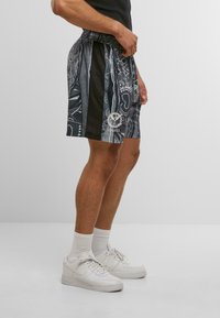 Sorte og grå shorts med et mønstret design, elastisk talje, sidelommer og et logo på venstre ben, parret med hvide sneakers.
