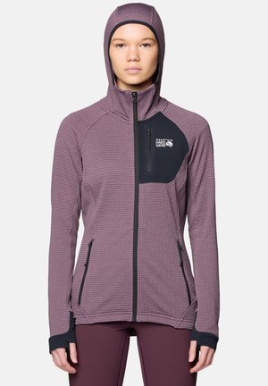 Femme portant une veste zippée texturée violette avec capuche et une poche poitrine noire, debout face à l'avant, les mains détendues le long du corps.