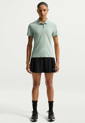 Frau steht und trägt ein mintgrünes Polo mit kurzen Ärmeln, einen schwarzen Sportrock, schwarze Socken und schwarze Sneakers vor einem einfarbigen Hintergrund.