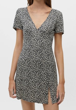 Robe noire à manches courtes avec motif floral blanc, encolure en V et fente sur le devant, portée par une femme debout aux cheveux bruns détachés.