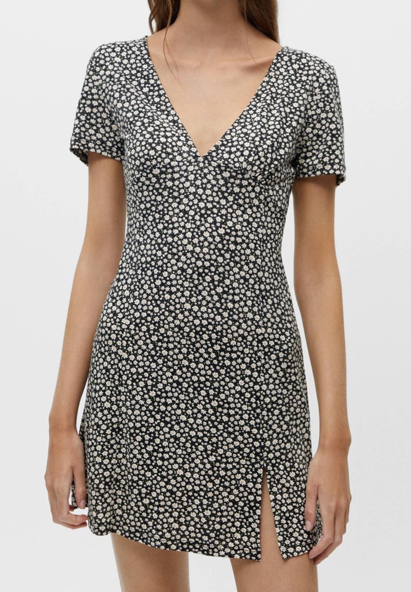 Robe noire à manches courtes avec motif floral blanc, encolure en V et fente sur le devant, portée par une femme debout aux cheveux bruns détachés.