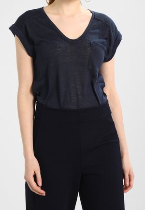Marineblå V-hals T-shirt med rullede korte ærmer, der har en lille brystlomme og en blød, let tekstur. Kombineret med mørke bukser.