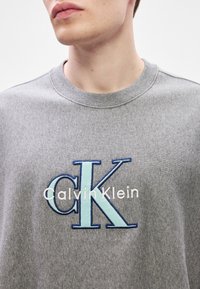 Šedá mikina s vyšívaným logem "CK" v modré a světle modré barvě, s nápisem "Calvin Klein" v bílé barvě pod ním. Měkká textura a kulatý výstřih.