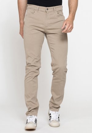 Uomo che indossa pantaloni beige slim fit e sneaker bianche con strisce nere, in piedi davanti a uno sfondo chiaro uniforme.
