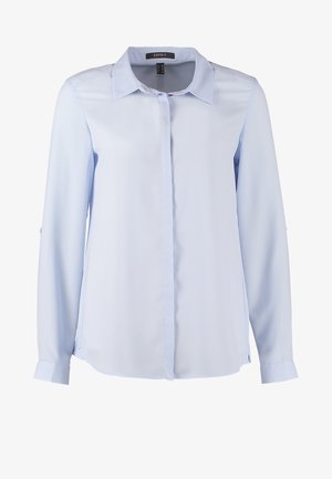 Lichtblauwe button-up blouse met opgerolde mouwen, gecombineerd met zwarte skinny jeans en zwarte hoge hakken. Het model houdt een zwarte clutch vast.
