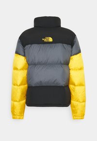 Kurtka puchowa z czarną górną częścią, szarą środkową i żółtą dolną sekcją. Posiada wysoki kołnierz i żółte logo „The North Face”.