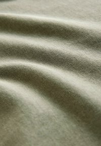 Olivovo zelená látka s hladkou textúrou, s jemnými vlnami a jemným tkaním. Tieňa vytvorené záhybmi zvýrazňujú hĺbku a detail.