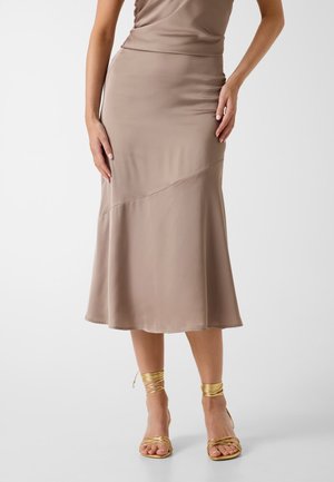 Femme portant une jupe midi taupe avec une couture asymétrique et des sandales à talons hauts métalliques dorées à lanières, debout devant un fond blanc.