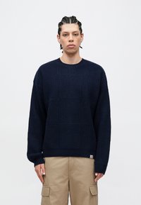 Maglione lavorato a maglia blu navy con un motivo testurizzato e decorato, collo rotondo e polsini a coste. Indossato con pantaloni cargo beige chiaro.