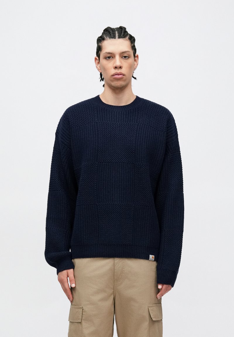 Navy blue knitted sweater featuring a textured, patterned design with a crew neckline and ribbed cuffs. Worn with light beige cargo pants.
