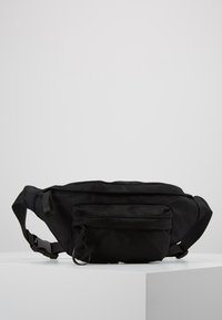 Riñonera negra texturizada con correa ajustable, que cuenta con un bolsillo delantero con cremallera y un diseño suave y redondeado. Material ligero.