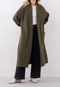Długi kardigan w oliwkowym kolorze z prążkowaną teksturą, otwarty z przodu, z dwiema przednimi kieszeniami i luźnym krojem, połączony z ciemnymi szerokimi spodniami i jasnymi loafersami.