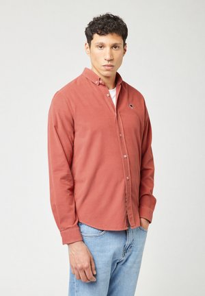 Roodbruine overhem shirt met lange mouwen, een opgerolde kraag en een klein logo detail. Ggedragen over een wit T-shirt, gecombineerd met blauwe jeans.