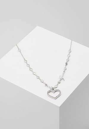 Collier en chaîne argentée avec de petits maillons en forme de cœur et un pendentif en forme de grand cœur ouvert incrusté de pierres claires, présenté sur une surface blanche.